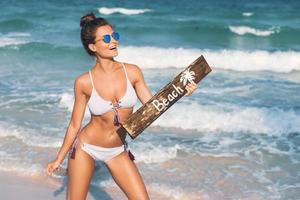 sexy donna indossare costume da bagno con vecchio di legno cartello su il spiaggia foto