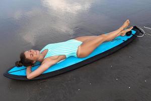 giovane donna indossare a strisce costume da bagno dire bugie su tavola da surf su il spiaggia foto