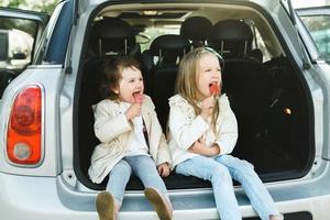 Due poco ragazze seduta nel un' macchine tronco e mangiare Lecca-lecca foto