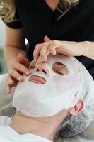 femmina cosmetologo l'applicazione foglio maschera su del cliente viso foto