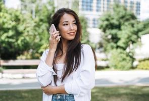 ritratto di giovane asiatico donna alunno con lungo capelli utilizzando mobile Telefono nel città parco foto