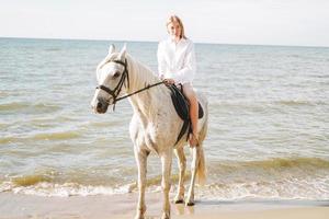 giovane rosso dai capelli donna nel bianca camicia equitazione bianca cavallo su paesaggio marino sfondo foto