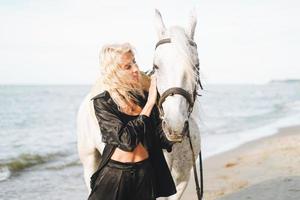 giovane bionda donna nel nero Abiti equitazione bianca cavallo su paesaggio marino sfondo foto