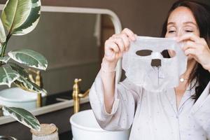 adulto brunetta donna con foglio maschera su sua viso nel il bagno a casa foto