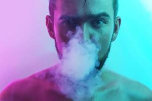 bello giovane uomo con bagnato pelle nel ultravioletto leggero fumo Vape foto