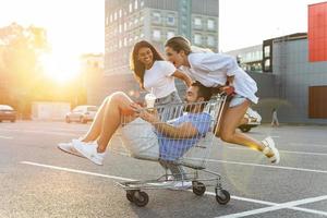 tre diverso amici avendo divertimento e equitazione shopping carrello su parcheggio lotto vicino supermercato foto