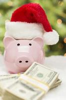 rosa porcellino banca indossare Santa cappello vicino pile di i soldi su i fiocchi di neve foto
