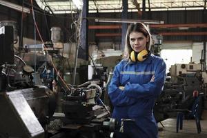 industriale lavoratore nel sicurezza uniforme con fiducioso braccia attraversato, giovane caucasico femmina ingegnere lavori con metallo macchine nel produzione fabbrica. professionale produzione meccanico occupazione. foto