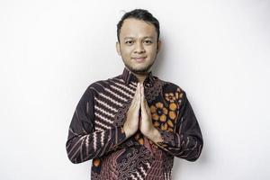 sorridente giovane asiatico uomo indossare batik camicia, Esprimere a gesti tradizionale saluto isolato al di sopra di bianca sfondo foto