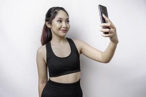 un' ritratto di un' sportivo asiatico donna indossare un' abbigliamento sportivo e Tenere sua Telefono, isolato di bianca sfondo foto