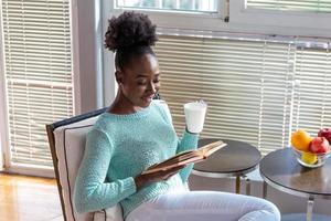 giovane donna con un' libro e tazza di caldo bere. bella afro americano ragazza con lettura un' libro seduta nel un' casa biblioteca con libreria nel il indietro e godendo sua caffè. foto