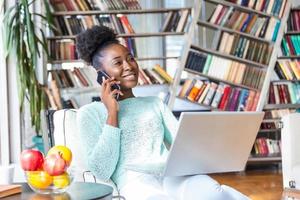 attraente giovane africano donna Lavorando su il computer portatile e sorridente mentre seduta su il divano a casa. bellissimo donna parlando su sua inteligente Telefono foto