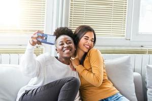 ridendo donna amici abbracciare ogni altro su divano mentre assunzione autoscatto foto su inteligente Telefono. amabile caucasico e africano americano ragazze esprimendo positivo emozioni per telecamera.