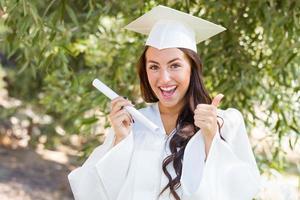 attraente misto gara ragazza festeggiare la laurea al di fuori nel berretto e toga con diploma nel mano foto