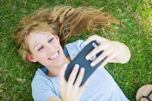 giovane adulto donna posa nel erba assunzione un' autoscatto con sua inteligente Telefono. foto
