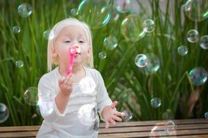 adorabile poco ragazza seduta su panchina avendo divertimento con soffiaggio bolle fuori. foto