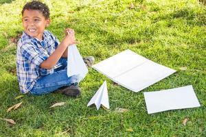 africano americano e messicano ragazzo apprendimento Come per piegare carta aeroplani all'aperto su il erba foto