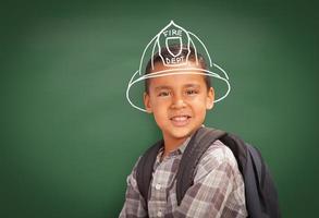 giovane ispanico alunno ragazzo indossare zaino davanti di lavagna con vigile del fuoco casco disegnato nel gesso al di sopra di testa foto