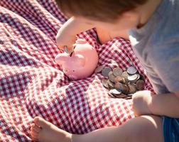 bambino ragazzo seduta su picnic coperta mettendo monete nel porcellino banca foto