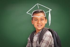 giovane ispanico alunno ragazzo indossare zaino davanti di lavagna con la laurea berretto disegnato nel gesso al di sopra di testa foto