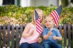 giovane sorella e fratello agitando americano bandiere su il panchina a il parco foto