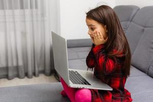 il poco ragazza si siede a un' tavolo con sorpresa sembra a il il computer portatile. su un' bianca sfondo. foto