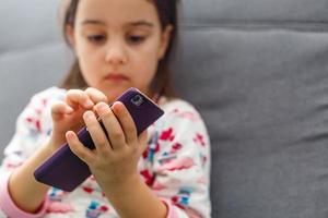 bellissimo poco ragazza giocando gioco o Guardando video su smartphone mobile. ragazza Guardando cartoni animati o navigazione Internet, copia spazio. lato Visualizza ritratto di poco ragazza utilizzando smartphone mentre seduta. foto