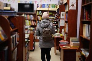 indietro di donna nel giacca e zaino raggiungendo un' libro a partire dal scaffale a il biblioteca. foto