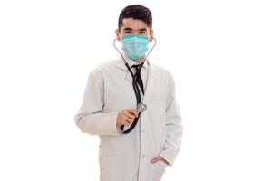 grave brunetta maschio medico nel uniforme con stetoscopio su il suo collo e maschera in posa e guardare a il telecamera isolato su bianca sfondo foto