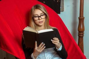 bellissimo ragazza indossare bicchieri con un' nero libro nel il suo mani su un' sedia foto