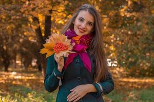 sorridente giovane ragazza sta nel autunno parco e mantiene il secco le foglie nel mano foto