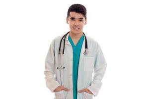 giovane bello brunetta uomo medico nel uniforme con stetoscopio su il suo le spalle guardare e sorridente su telecamera isolato su bianca sfondo foto