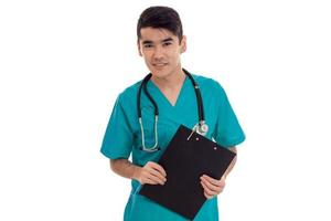 giovane medico nel un' blu grembiule con stetoscopio su il le spalle Tenere un' nero tavoletta per record e sorridente isolato foto
