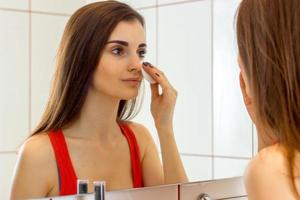 bellissimo giovane ragazza nel un' rosso camicia sta nel davanti di un' specchio e salviette il viso con un' cotone disco foto