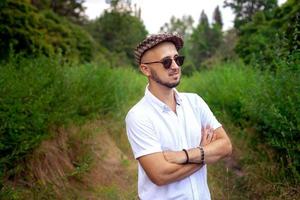 fascino giovane uomo modello nel cappello detiene occhiali da sole e guardare lontano all'aperto foto