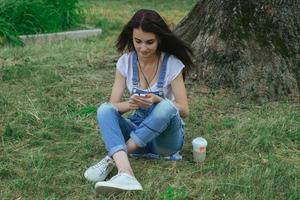 giovane affascinante brunetta si siede nel il erba ascoltando per musica e guardare a Telefono foto