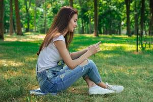 affascinante ragazza si siede nel il parco su il erba e guardare a il Telefono foto