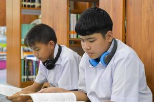 morbido fucus di Due asiatico ragazzo studenti siamo ascoltando media, lettura e consulenza di preferito libro a biblioteca di scuola foto