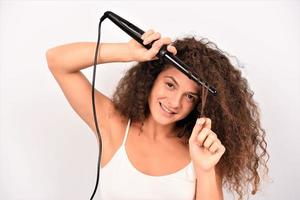 Riccio capelli. bellissimo sorridente donna con lungo ondulato capelli Stirare esso, utilizzando arricciatura ferro da stiro. contento ragazza con bellissima salutare liscio capelli utilizzando bigodino per Perfetto riccioli. acconciatura e parrucchiere foto