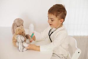 carino poco ragazzo giocando medico a casa e curare felpa giocattolo. dolce bambino piccolo bambino giochi interno. avendo divertimento. bambini e medicinale, assistenza sanitaria. foto