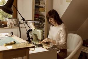 caucasico brunetta donna cucire tessuto con un' Vintage ▾, retrò cucire macchina. moda, creazione e sartoria. processi di cucire nel Atelier o officina. speciale passatempo. accogliente interno e caldo colori. foto