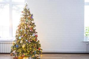 Natale albero nel il bianca interno di un' Casa con grande finestre. raggiante Fata luci ghirlande interno decorazione di il studio camera. in vaso impianti nel il casa foto
