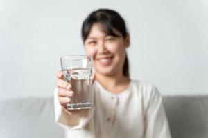 salutare bellissimo giovane donna Tenere un' bicchiere di acqua seduta su il divano a il soggiorno. foto