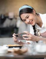 giovane donna indossa grembiule assunzione autoscatto foto con fatti in casa torta nel cucina. ritratto di bellissimo asiatico femmina cottura al forno dolce e avendo divertimento assunzione foto di smartphone per in linea sociale. casa cucina.