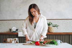 attraente adulto mezzo anziano brunetta donna cinquanta anni nel bianca camicia cucinando insalata di fresco verdure nel cucina a il casa foto