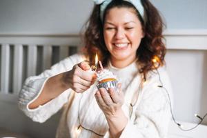 giovane adulto contento bellissimo donna quaranta anni più dimensione corpo positivo nel bianca accogliente a maglia maglione con festivo Cupcake con candela nel mani su letto a casa festa, compleanno ragazza foto