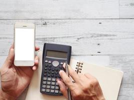 mano Tenere mobile Telefono bianca schermo e utilizzando calcolatrice con cactus e argento penna su bianca legna tavolo natura ombra e luce del sole superiore Visualizza e spazio attività commerciale concetto foto