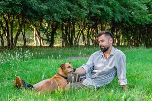 bello uomo nel occhiali da sole seduta su erba con il suo cane nel parco. concetto di umano e animale domestico relazione. foto