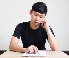 asiatico uomo sedersi a il scrivania dai un'occhiata elenco su tavola e parlando mobile Telefono grave viso foto