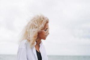 ritratto di elegante bionda donna nel bianca camicia su sabbia spiaggia a tempesta mare a ventoso weater foto